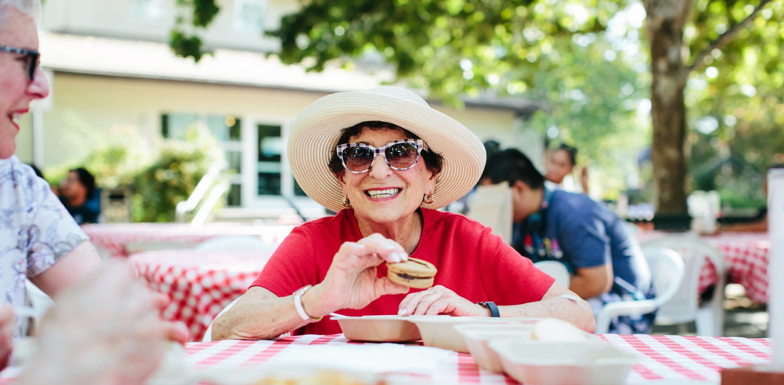 A smiling older woman wearing sunglasses and a hat eats a snack at an outdoor picnic table